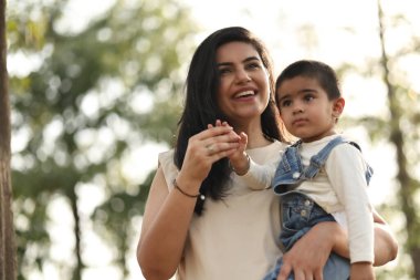 Gülümseyen bir anne, çocuğunu ağaçlarla çevrili güneşli bir parkta tutar..
