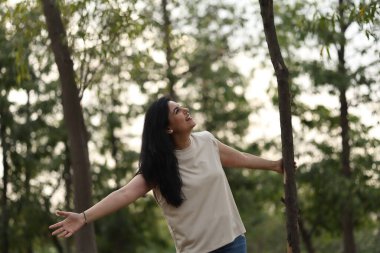 Gün batımında ağaçlarla çevrili sakin bir parkta kadın gülümser ve doğayı kucaklar..