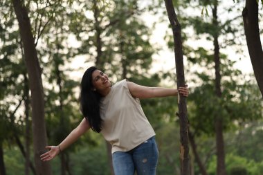 Bir kadın, akşam vakti parkta bir ağacın yanında durmuş, dışarıyı kucaklıyor..
