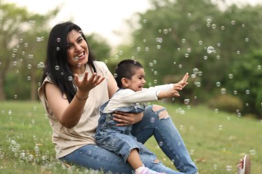 Neşeli bir anne ve küçük oğlu, çimenli bir parkta yüzen baloncuklara uzanarak birlikte oynuyorlar..