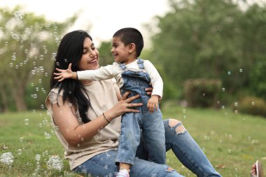 Anne ve küçük çocuk güneşli bir parkta baloncuklarla oynarken keyifli anları paylaşıyorlar..