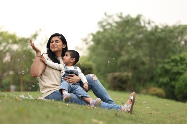 Anne ve çocuk güneşli bir günde yeşil çimlerde kabarcıklarla oynarken neşeli anları paylaşırlar..