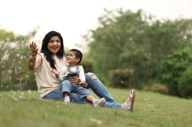 Bir anne ve küçük oğlu yeşil bir parkta baloncukları üflemekten ve yakalamaktan zevk alırlar..