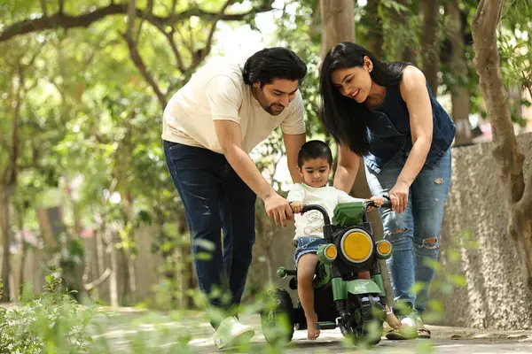 Ebeveynler, yeşil alanlarla çevrili bir yolda oyuncak bir araç kullanan küçük çocuklarına rehberlik ederler..