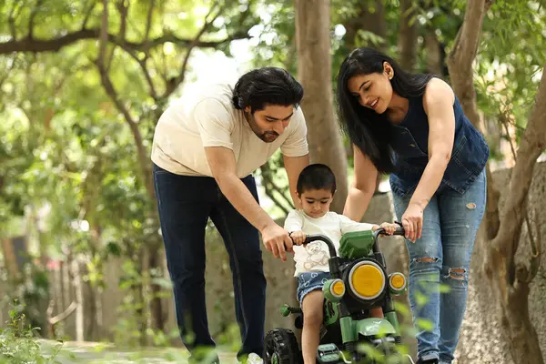 Ebeveynler, o yeşil, güneşli parkta oyuncak bir araca binerken çocuklarına rehberlik ederler..