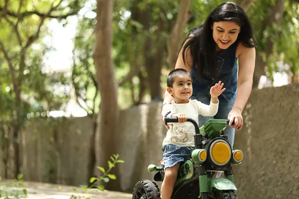 Bir kadın, yemyeşil bir park ortamında çocuğunun küçük bir motorsiklet sürmesine yardım ediyor..