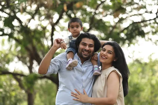 Bir adam omuzlarında küçük bir çocuk taşırken bir kadın yemyeşil bir parkta onların yanında gülümsüyor..