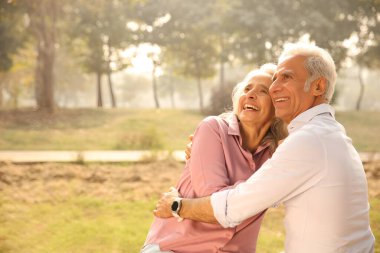Mutlu çift, güneşli bir parkta sıcak bir kucaklaşmayı ve kahkahayı paylaşıyor. Altın yıllarını mutlulukla kucaklıyorlar..