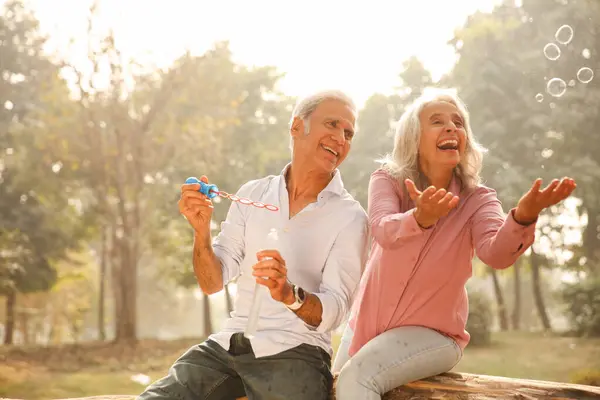 Yaşlı bir yetişkin, güneşli bir park ortamında baloncuklarla oynar, güler ve eğlenir..