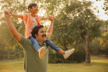 Baba parkta oğlunu omuzlarında taşırken neşeli bir oyun ve bağ kurma anını paylaşıyor..