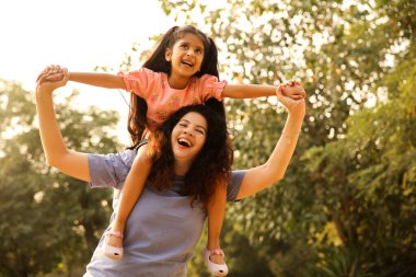 Anne kızını omuzlarında taşıyor, hem gülüyor hem de parkta eğleniyor..