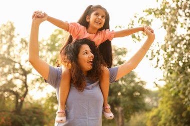 Anne ve kızı parkta oynuyorlar, kız annesinin omuzlarında neşeyle ata biniyor..