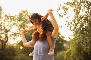 Anne ve kızı neşeli bir anın tadını çıkarıyorlar, kız annesinin omuzlarında, sakin bir açık hava ortamında.