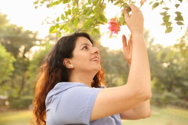 Güzel bir kadın gür bir bahçede canlı kırmızı çiçeklere dokunuyor, dinginlik ve zarafet saçıyor..