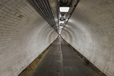 Beyaz kiremitli duvarları ve tepeden aydınlatması olan uzun bir yeraltı tünelinin perspektif görüntüsü. Endüstriyel borular tavanda akıyor, minimalist estetiği güçlendiriyor..