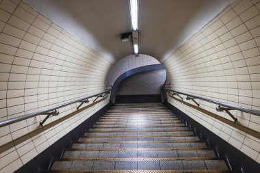 Loş bir yeraltı metro tüneli fayanslı merdiven, metal korkuluklar ve florasan aydınlatma içeren karamsar ve simetrik bir perspektif yaratıyor.