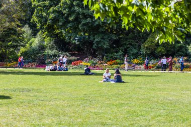 Canlı çiçekler ve güneş ışığında ağaçlarla çevrili yemyeşil çimlerde dinlenen insanların olduğu sakin bir park sahnesi.