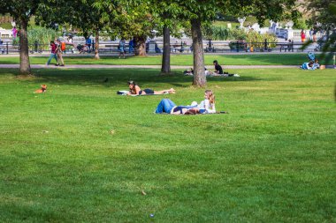 Şehir parkında güneşli bir günün tadını çıkaran ziyaretçiler ağaçlar ve açık alanlarla çevrili yeşil çimlerde dinleniyorlar.