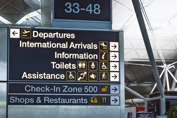 Gidiş yönlerini, geliş yönlerini ve yolcu hizmetlerini gösteren bir havaalanı tabelasını kapatın. Londra, İngiltere, 18 Kasım 2023