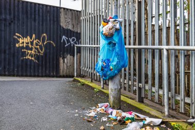 Metal çitlere asılı mavi plastik çöp torbası ve etrafa saçılmış çöp..
