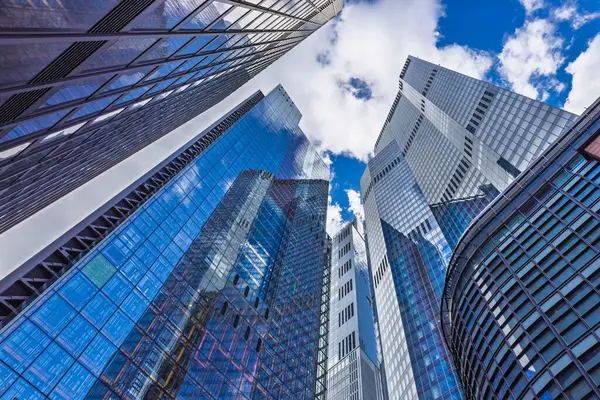 Çağdaş gökdelenlerin göz kamaştırıcı manzarası yansıtıcı cam cepheleriyle parlak mavi gökyüzü altında bulutlarla.