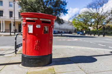 Geleneksel kırmızı İngiliz posta kutusunu mavi gökyüzü ve yapraklı ağaçlarla dolu bir kaldırımda kapatın. Londra, İngiltere, 1 Nisan 2024