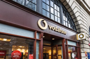 Vodafone perakende mağazasının dış tarafında ışıklı logo işareti ve şehir ortamında büyük cam pencereler var. Londra, İngiltere, 1 Nisan 2024