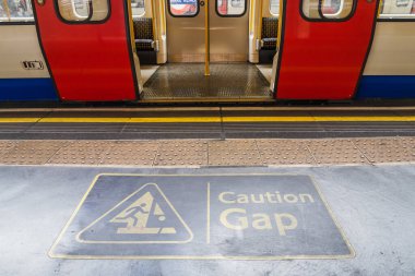 Londra yeraltı platformunda CAUTION GAP güvenlik levhası, görünür tren kapıları.