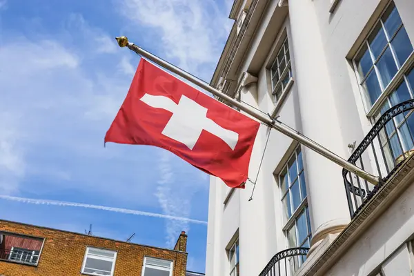 İsviçre bayrağı, mavi gökyüzüne karşı canlı kırmızı ve beyaz bir haç dalgalandırıyor..