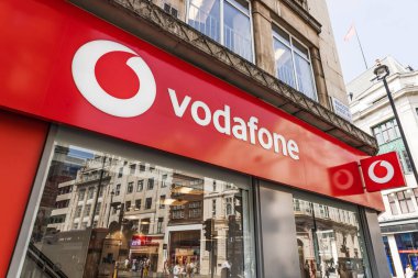 Londra şehir merkezindeki Vodafone perakende mağazasının geniş açılı çekimi, güneşli kentsel koşullar altında koyu kırmızı markalaşma, cam cephe ve sokak yansımaları içeriyor. Londra, İngiltere, 11 Mayıs 2024