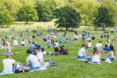 Parlak bir yaz öğleden sonrasında piknik yapan, sohbet eden ve güneşlenen insanlarla dolu canlı bir şehir parkının geniş panoramik manzarası. Londra, İngiltere, 2 Haziran 2024