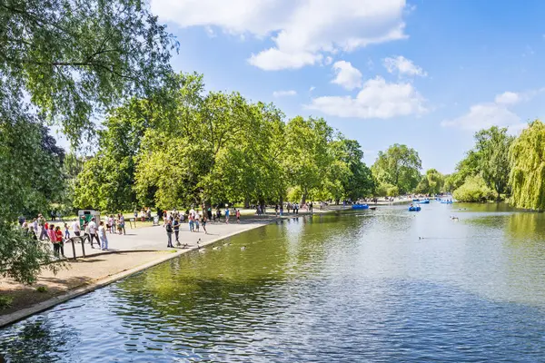 Panoramik Vekiller Park Tekne Gölü, Londra. İnsanlar güneşli yolda yürür, kayıklar sakin sularda süzülür, yemyeşil ağaçlar ve mavi gökyüzü cennet gibi bir yaz şehri kaçışı yaratır. Londra, İngiltere, 2 Haziran 2024