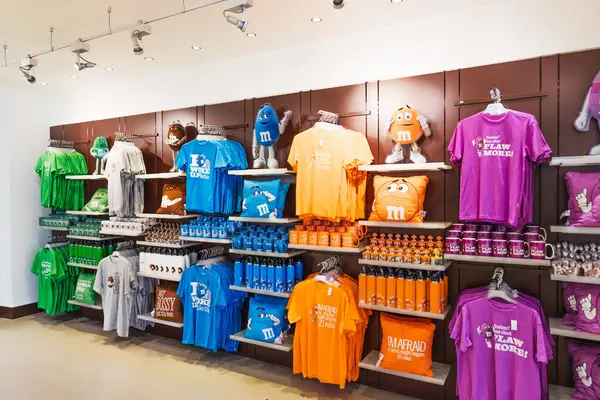 Organize M ve M ticaret duvarı. Gökkuşağı tişörtleri, kapüşonlular, kupalar ve hediye kutuları parlak şeker dükkanında düzgünce istiflenmiş, perakende satışı ve renkli kıyafetler sergileniyor. Londra, İngiltere, 2 Haziran 2024