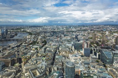 Londra 'nın yüksek açılı panoramasında Thames Nehri yoğun bloklar ve kuleler arasında dolanıyor. Geniş şehir silueti, dramatik bulutlar, iş bölgesi ve simgeler görünür. Londra, İngiltere, 7 Temmuz 2024