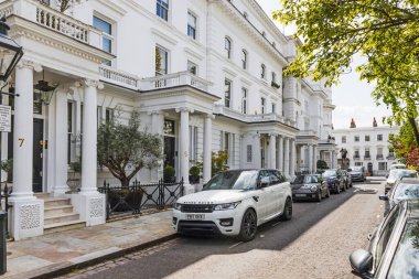 Sakin bir Londra caddesinde, park etmiş arabaları, verandaları ve balkonları olan zarif, beyaz teraslı evler. Londra, İngiltere, 24 Mayıs 2023