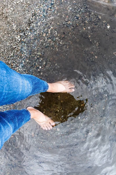 Woman's legs in jeans with bare soles standing in water.