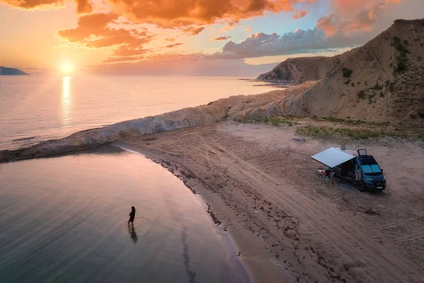 Independent road trip concept: Campervan on Abandoned Beach against beutiful Sunset,  Woman bathing in the Sparkling sea. Outdoor nomad lifestyle, van life holiday. Aerial photo. Albania coast.