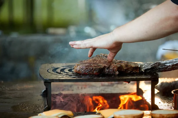 A chef controls a steak on an outdoor grill by finger. Close up of cooking at garden festival, flames, shallow depth of field, very colorful blurred background, evening at garden festival.