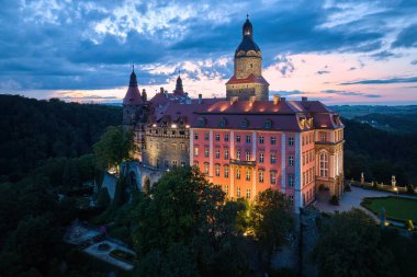Panoramik, aydınlanmış Ksiaz Şatosu 'nun akşam manzarası, Schloss Frstenstein, dramatik bir gökyüzüne karşı ormanla çevrili bir kayanın üzerinde duran güzel bir kale. Polonya