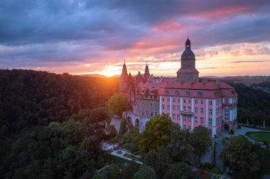 Panoramik, kırmızı güneşli Ksiaz Kalesi manzaralı, Schloss Frstenstein, dramatik bir gökyüzüne karşı ormanla çevrili bir kayanın üzerinde duran güzel bir kale. Polonya