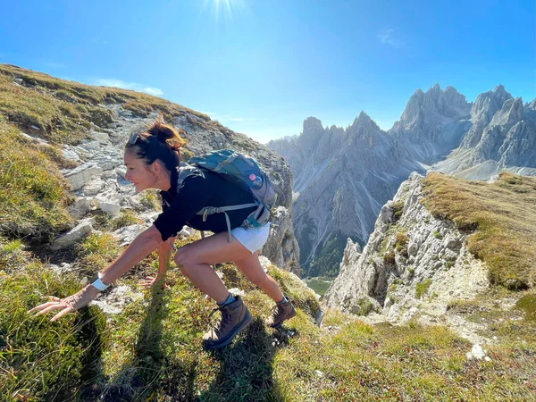 Sporting at an older age theme: sporty older woman on a trek in the Dolomites, red shirt, shorts, dark hair, backpack, sunny day. Physically demanding climb, rocks, mountains and forests in the back