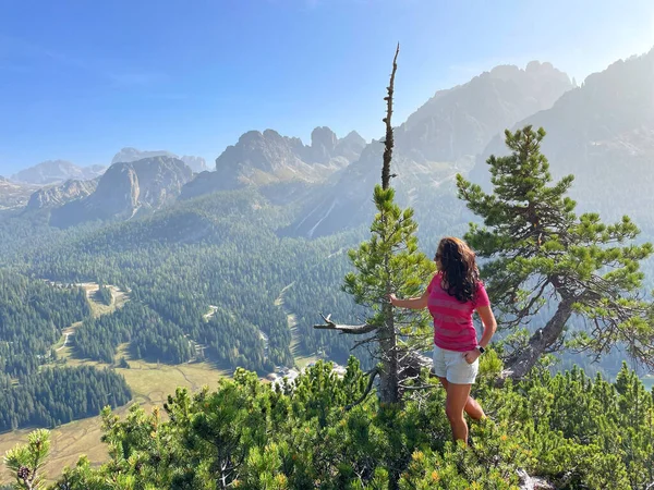 Freshly retired theme: sporty older woman on a trek in the Dolomites, red t-shirt, shorts, dark hair, backpack. Physically demanding climb, mountains and forests in the background.