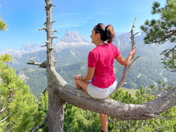 Freshly retired theme: sporty older woman on a trek in the Dolomites, red t-shirt, shorts, dark hair, backpack. Physically demanding climb, mountains and forests in the background.