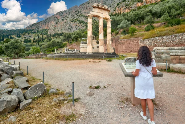 白いドレスの長髪の女性は デルファイのアテナ プロナイアの古代神殿複合体のボードから情報を読み取ります 晴れた日 ユネスコ世界遺産 ロイヤリティフリーのストック写真
