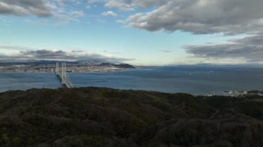 Stationary aerial view of suspension bridge spanning wide straight to distant city on partly cloudy afternoon. 