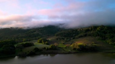 Pembe sis ve benekli sabah güneşi, şafak vakti gölün yanındaki yeşil tepelerde. Yüksek kalite 4k görüntü