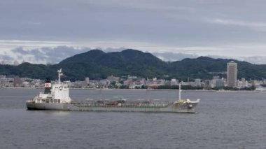 Kobe, Japonya - 23 Eylül 2023: kıyı kasabası ve dağlardan geçen küçük bir kargo gemisiyle denize açılmak. Yüksek kalite 4k görüntü