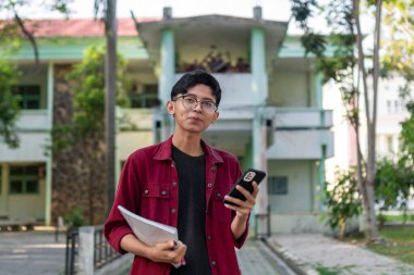 Genç Asyalı üniversite öğrencisi mutlu bir ifadeyle akıllı telefon kullanıyor. Halk parkında elinde telefon ve kitaplarla gülümseyen bir erkek. Boşluğu kopyala