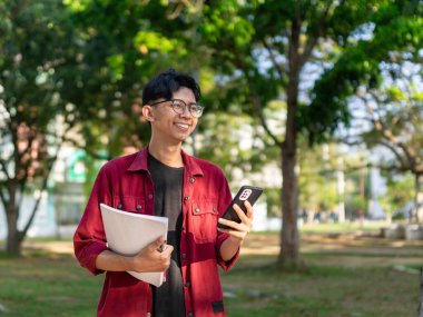 Genç Asyalı üniversite öğrencisi mutlu bir ifadeyle akıllı telefon kullanıyor. Halk parkında elinde telefon ve kitaplarla gülümseyen bir erkek. Boşluğu kopyala