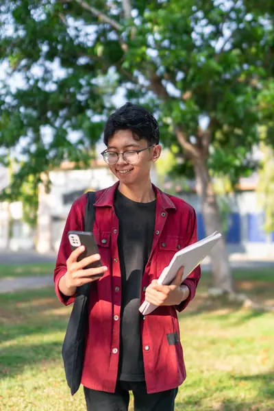 ハッピーな表情でスマートフォンを使用したアジアの若い大学生 公立公園で携帯と本を持っている間 男性は笑顔で コピースペース — ストック写真